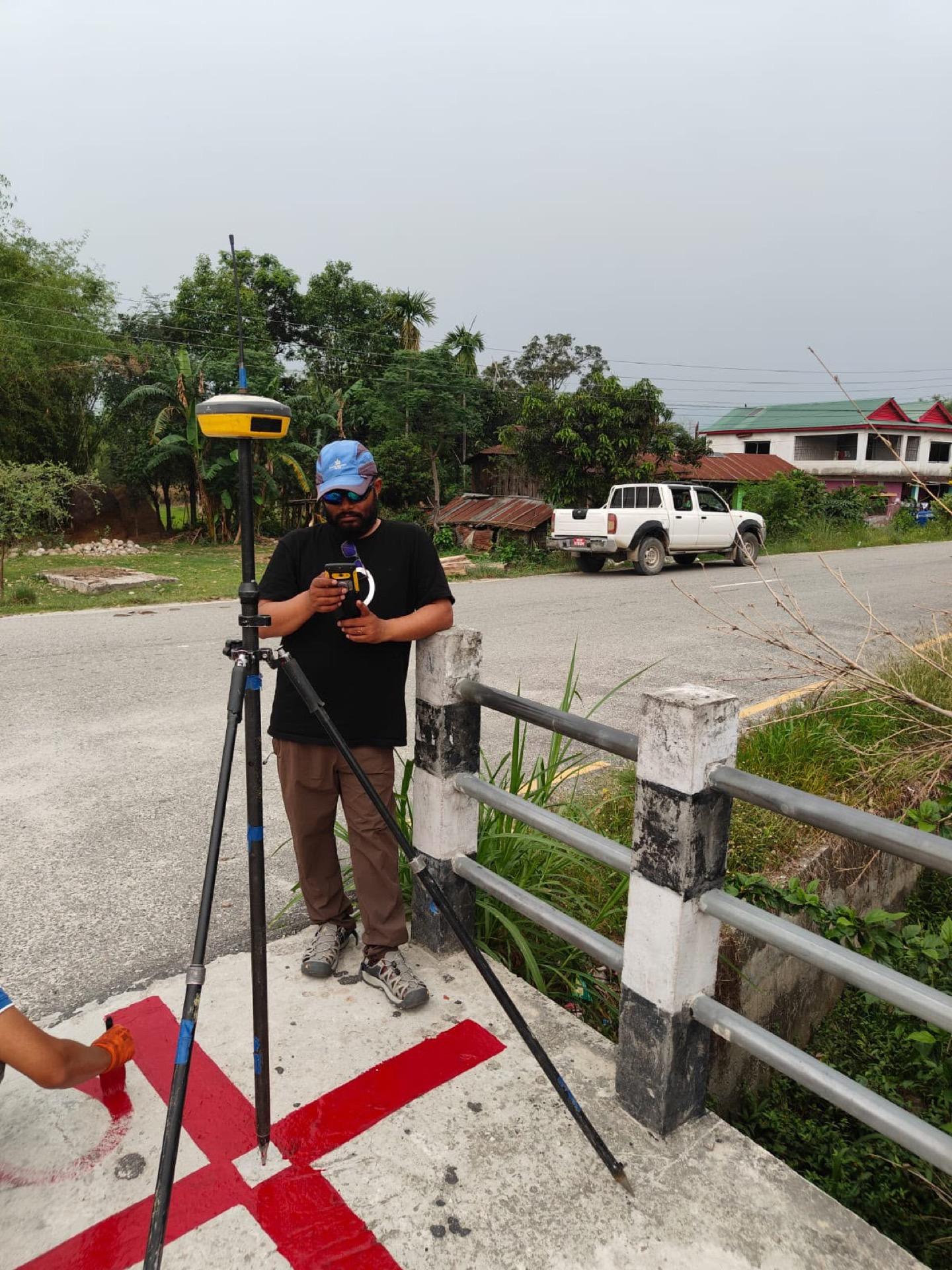 GNSS receiver mounted on a tripod over a painted survey benchmark beside a bridge